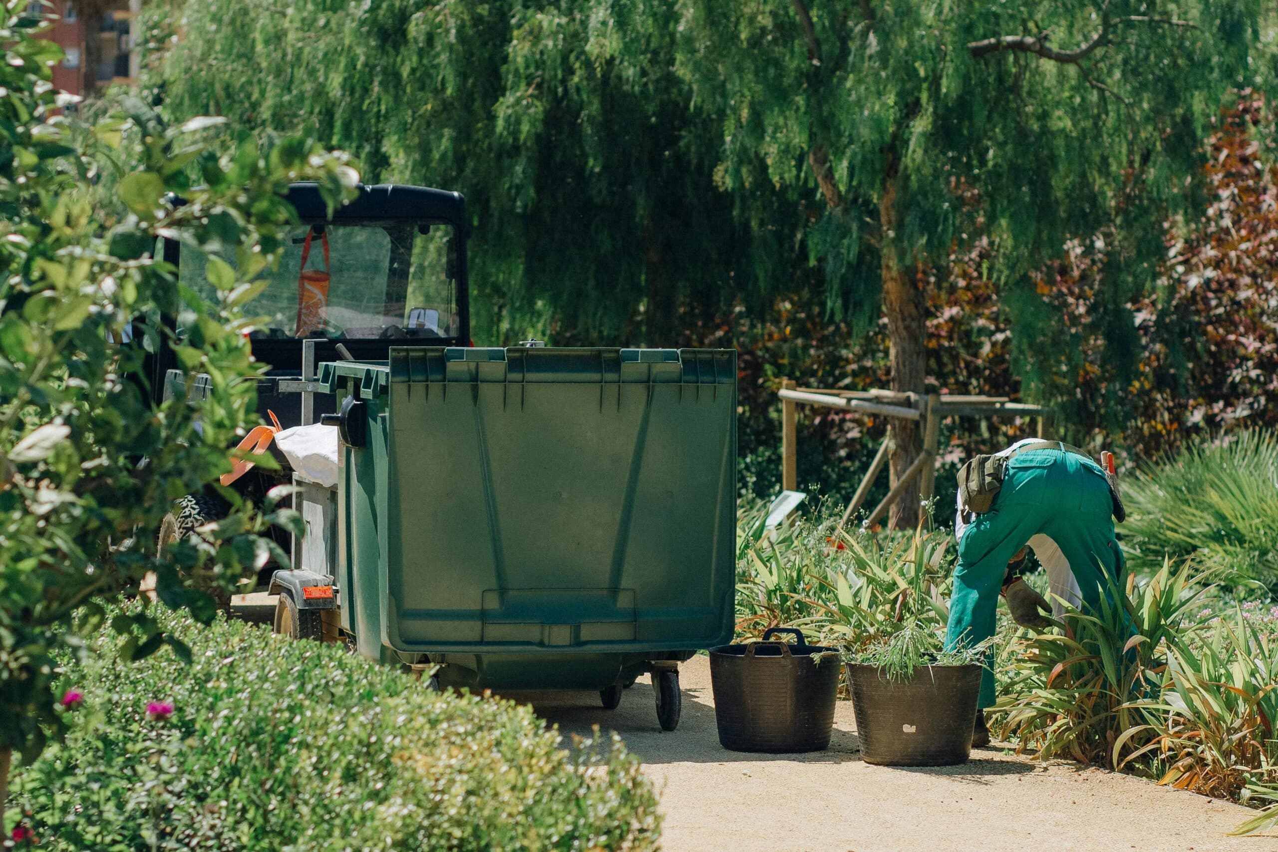 A landscaper in green work clothes tends to plants in a lush Fairfield, California garden near a utility vehicle and large bin, possibly representing one of the best landscaping companies in Fairfield.